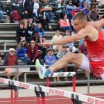 A male athlete in red clears a hurdle during a track event.